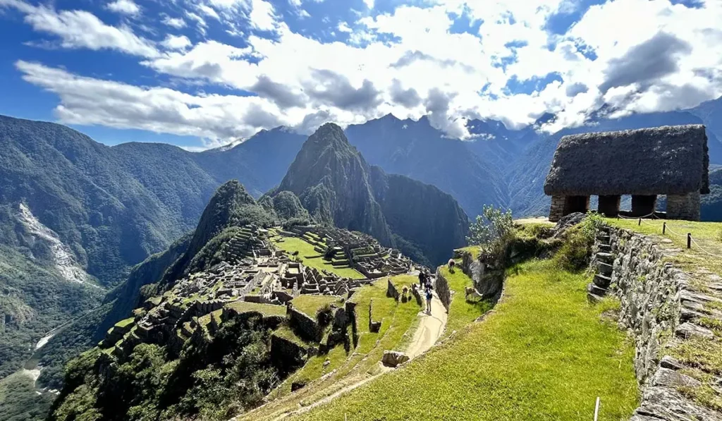 Vista panorámica de Machu Picchu bajo cielo azul con nubes blancas. Se aprecia la Casa del Guardián, terrazas incas y las montañas Huayna Picchu y Putucusi. Sitio arqueológico Inca en Cusco, Perú.